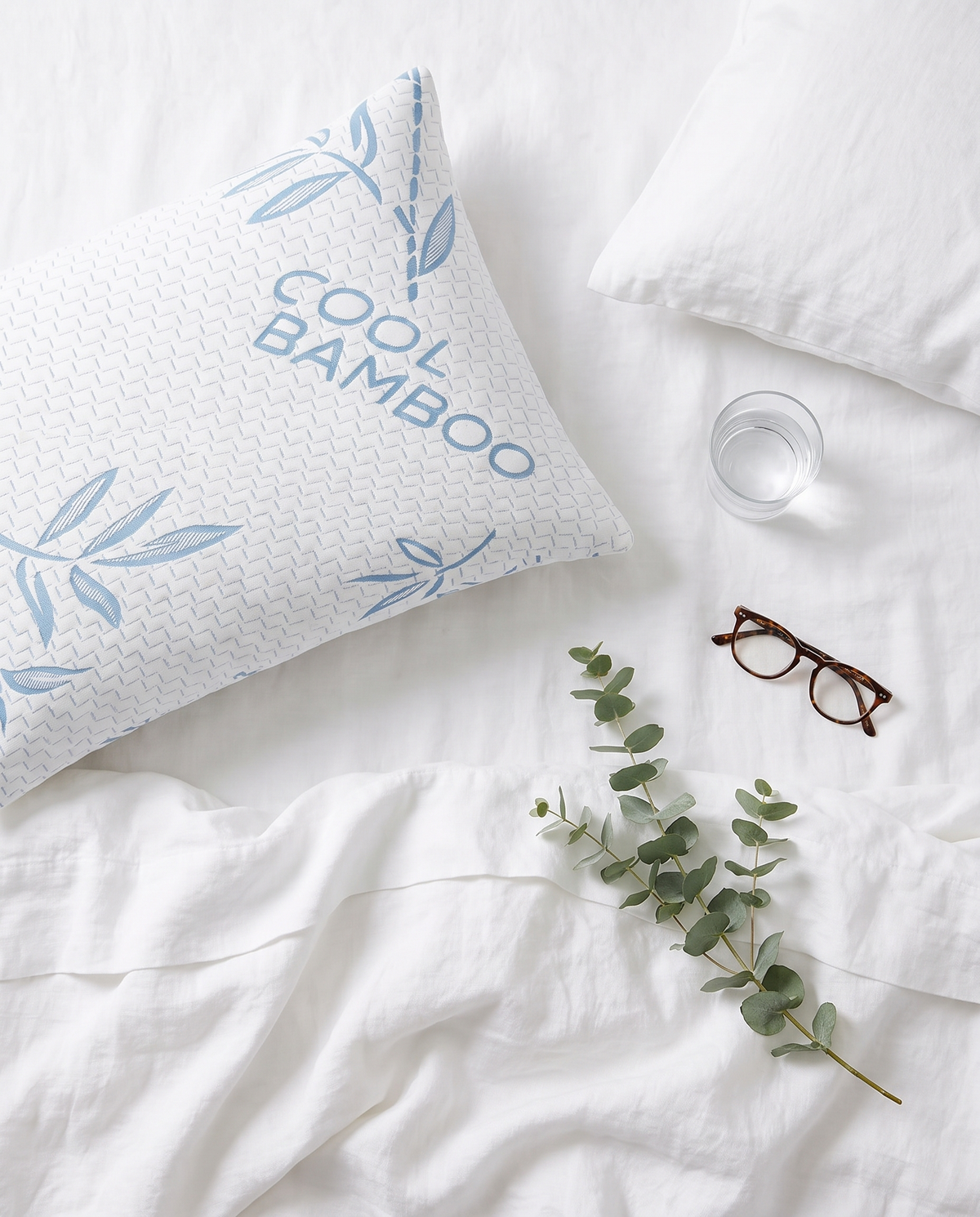 Top-down view of an OMEO blue bamboo pillow on white sheets with a glass of water, reading glasses and eucalyptus