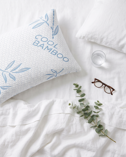 Top-down view of an OMEO blue bamboo pillow on white sheets with a glass of water, reading glasses and eucalyptus